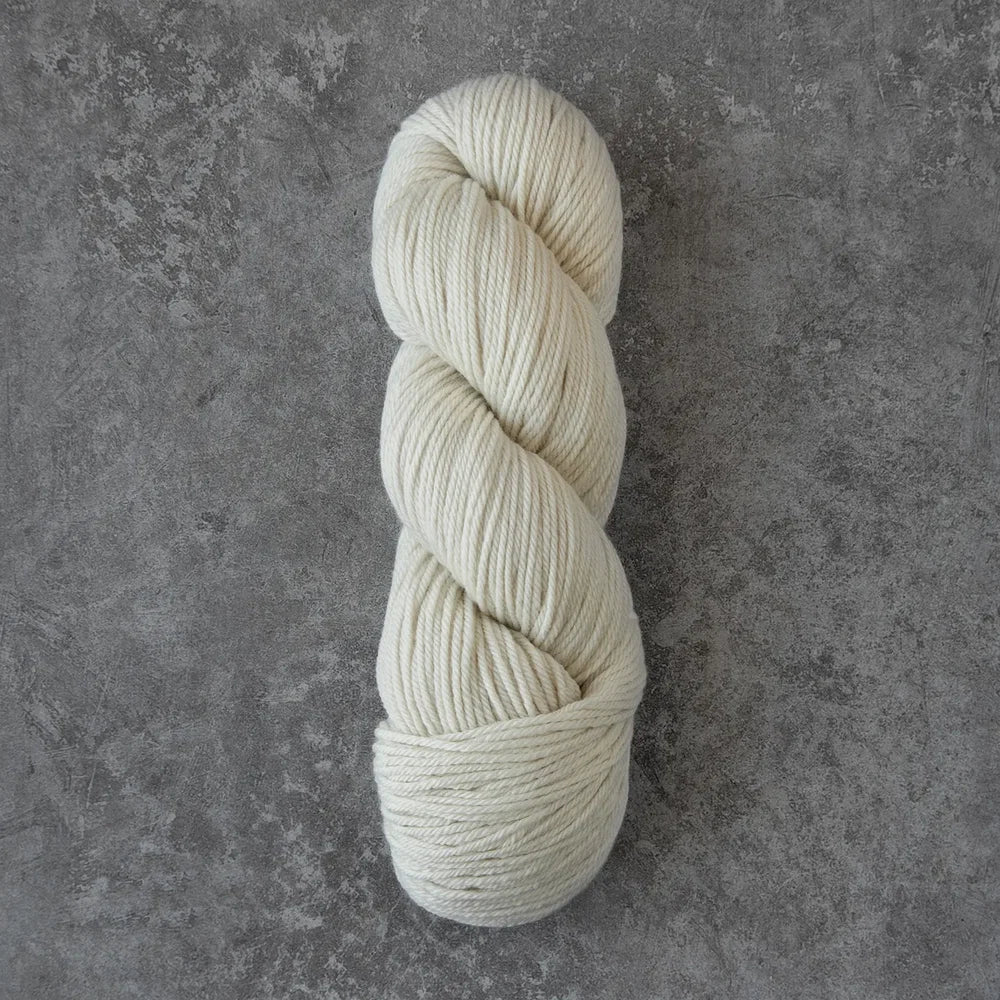 A single skein of cream-colored yarn twisted into a balm, displayed vertically on a textured gray concrete surface.