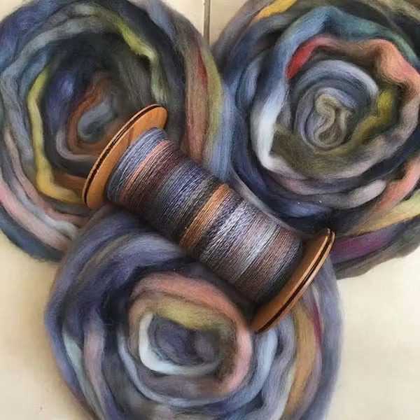 Three rosettes of soft, multi-colored wool roving with a wooden spool threaded with matching variegated wool, set against a light beige background.
