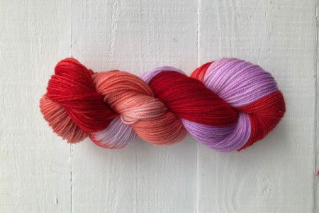 A twisted hank of variegated yarn with a color gradient from bright red to coral, peach, and lavender purple, displayed on a white wooden background.