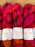 Three skeins of Barni New York 100% Superwash Merino Wool yarn in a variegated red, orange, and pink colorway, displayed on a wooden surface with white paper labels.