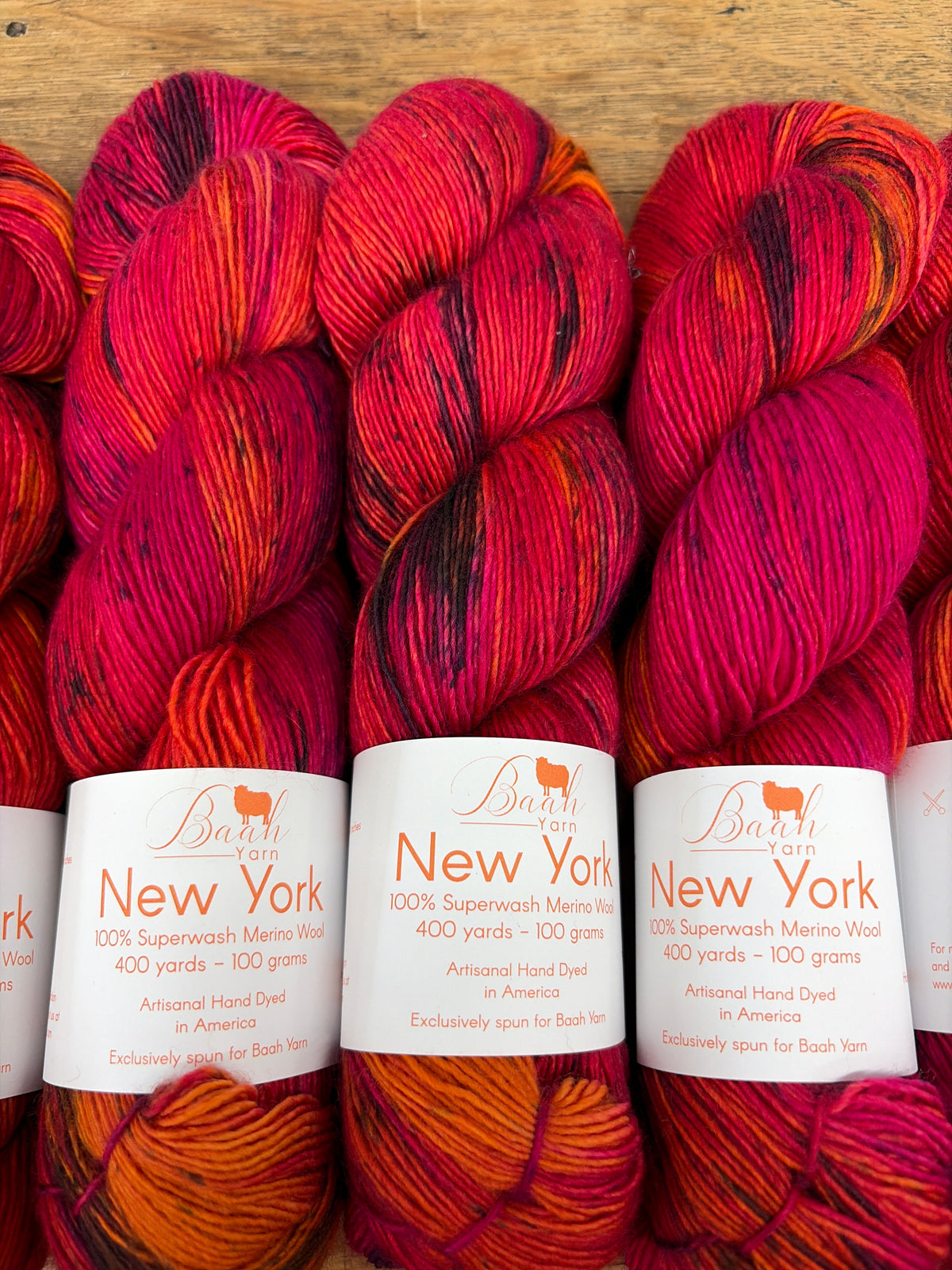 Three skeins of Barni New York 100% Superwash Merino Wool yarn in a variegated red, orange, and pink colorway, displayed on a wooden surface with white paper labels.