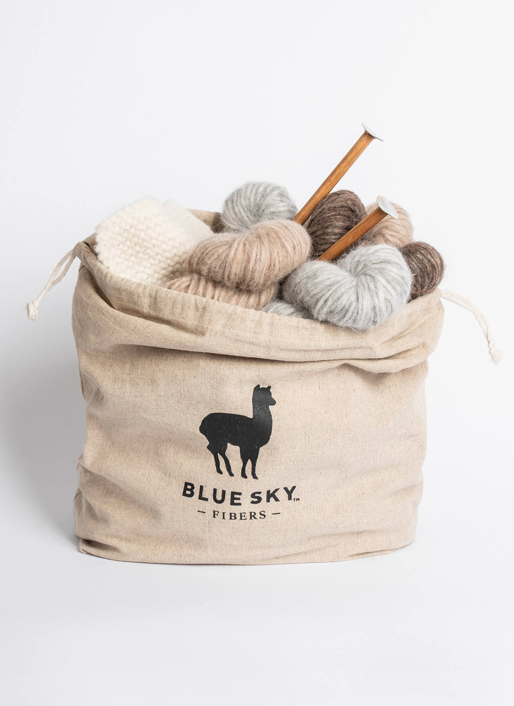 A beige canvas drawstring bag with a black alpaca logo and the text 'BLUE SKY. FIBERS' on the front, filled with balls of yarn in cream, tan, brown, and light gray, with two wooden knitting needles sticking out, on a white background.