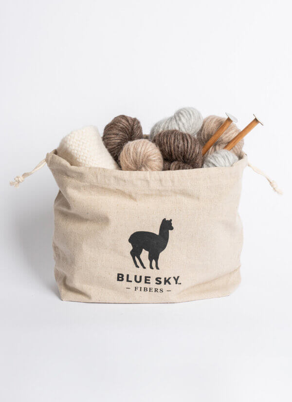 A beige canvas drawstring bag with a black alpaca silhouette logo and the text 'BLUE SKY. FIBERS' printed on the front, filled with balls of yarn and wooden knitting needles against a white background.