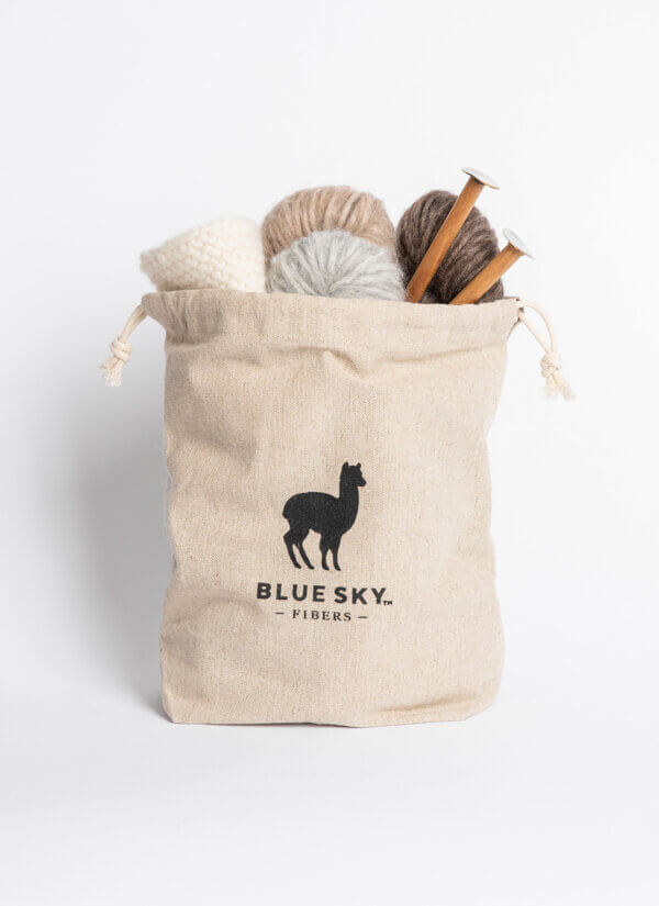A beige canvas drawstring bag with a black alpaca silhouette logo and the text 'BLUE SKY. FIBERS.