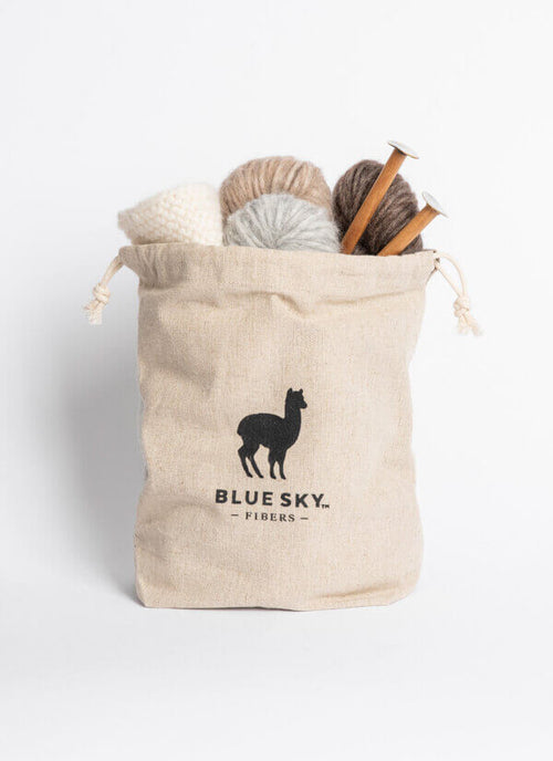 A beige canvas drawstring bag with a black alpaca silhouette logo and the text 'BLUE SKY. FIBERS.