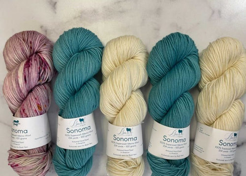 Five skeins of Sonoma 100% Superfine Merino wool yarn in assorted colors, including variegated pink and cream, teal, and solid cream, arranged on a light marble surface.