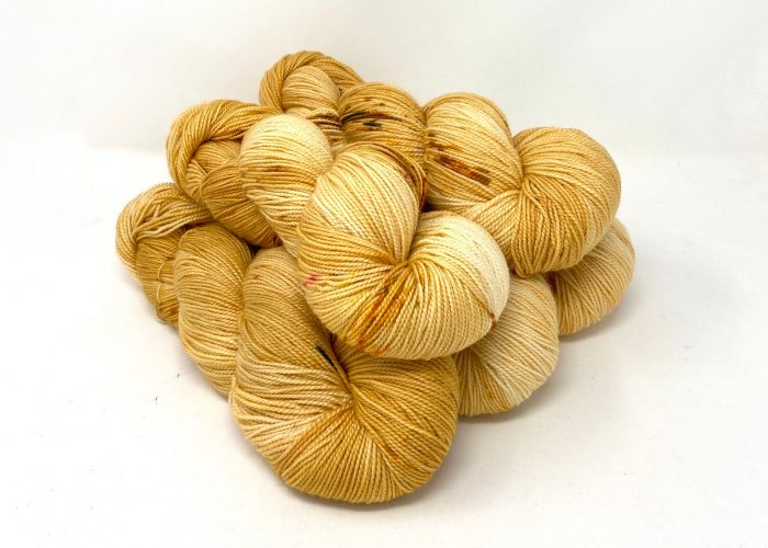 A pile of hand-dyed yarn skeins in a variegated blend of pale cream, light yellow, and golden ochre on a white background.