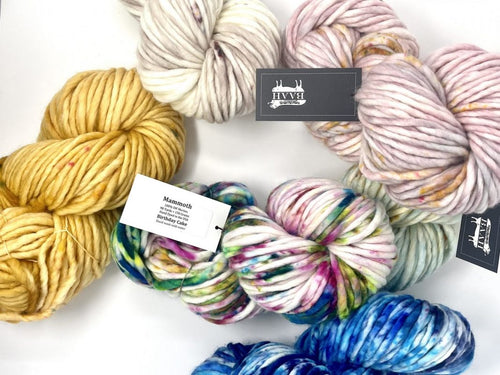 A flat-lay arrangement of multiple skeins of variegated yarn on a white background, featuring brands 'Knit With Love' and 'Mammoth'. The yarns display various colorways including solid mustard yellow, cream, dusty pink, and variegated patterns of blue, green, purple, and white.