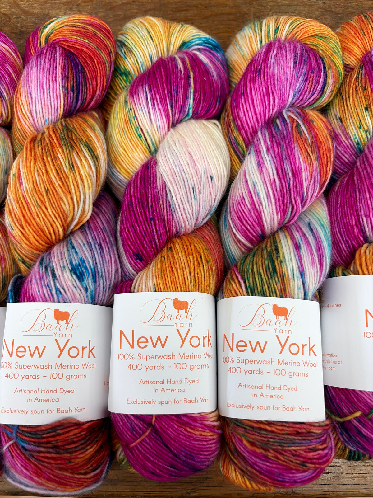 Five skeins of variegated yarn from Bash Yarn's New York series arranged in a row on a wooden surface, showcasing vibrant multicolor blends of orange, pink, purple, yellow, and blue.