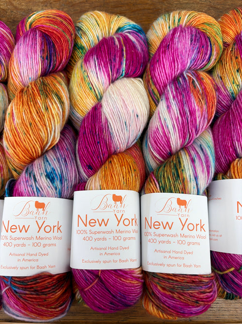 Five skeins of variegated yarn from Bash Yarn's New York series arranged in a row on a wooden surface, showcasing vibrant multicolor blends of orange, pink, purple, yellow, and blue.