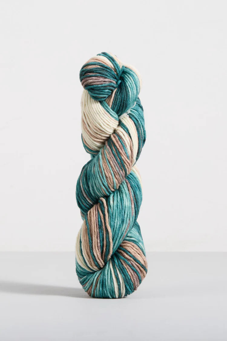 A single twisted hank of variegated yarn standing upright on a light gray surface against a white background. The yarn has a variegated colorway of teal, cream, and dusty pink.