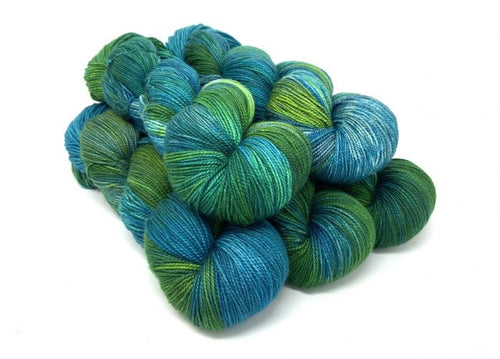 A pile of hand-dyed yarn skeins in a variegated blend of teal, blue, and green, photographed on a clean white background.