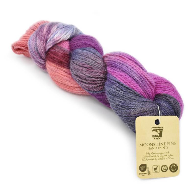 A skein of Moonshine Fine Hand Paint yarn from Prairie Dawn Yarn, featuring a variegated color pattern of pink, purple, and blue, with its paper tag attached, on a white background.