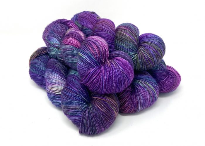 A pile of hand-dyed fingering yarn skeins in a variegated blend of purple, blue, and pink, photographed on a clean white background.