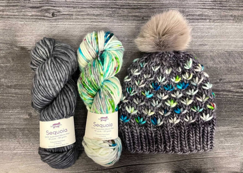 A handmade knit beanie with a large beige pom-pom, shown alongside two skeins of yarn on a gray wood background. The beanie is made from a dark gray and white variegated yarn with blue, green, and teal leaf-like accents.