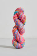 A single twisted hank of variegated yarn standing upright on a light gray studio background. The yarn shows multiple bright colors including pink, blue, teal, yellow, and orange in roughly equal prominence.