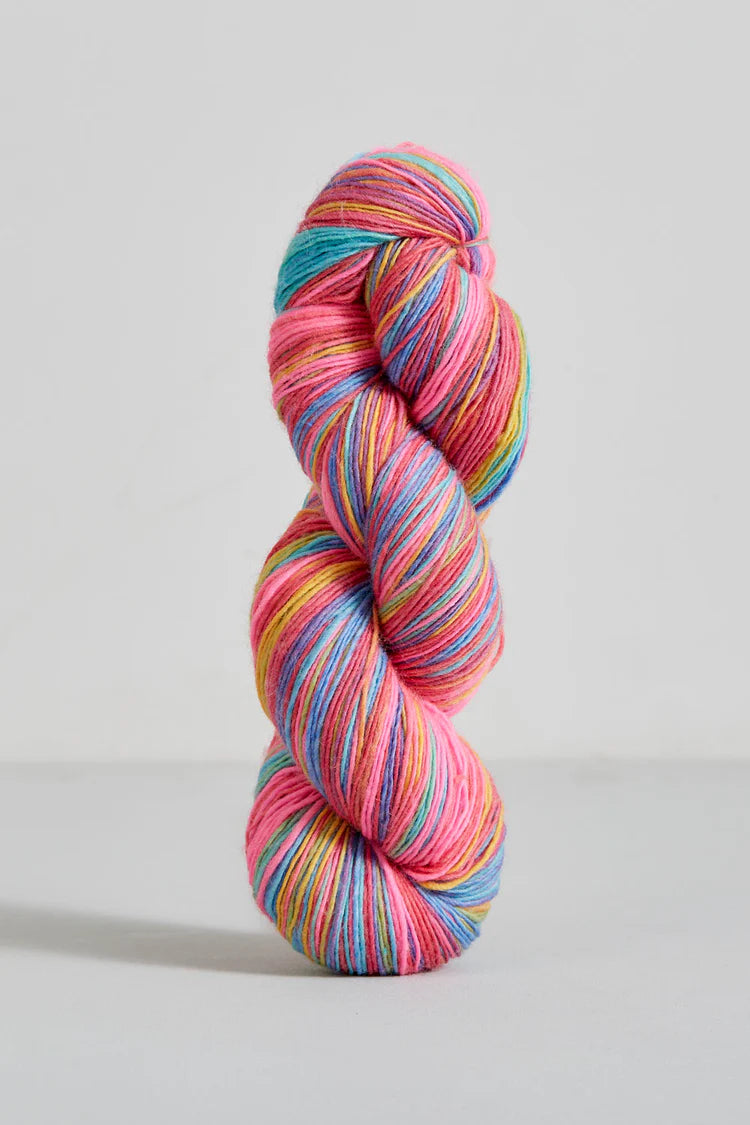 A single twisted hank of variegated yarn standing upright on a light gray studio background. The yarn shows multiple bright colors including pink, blue, teal, yellow, and orange in roughly equal prominence.