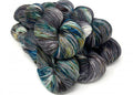 A pile of hand-dyed hanks of yarn with variegated colors of charcoal gray, navy, teal, olive green, and white, arranged on a plain white background.