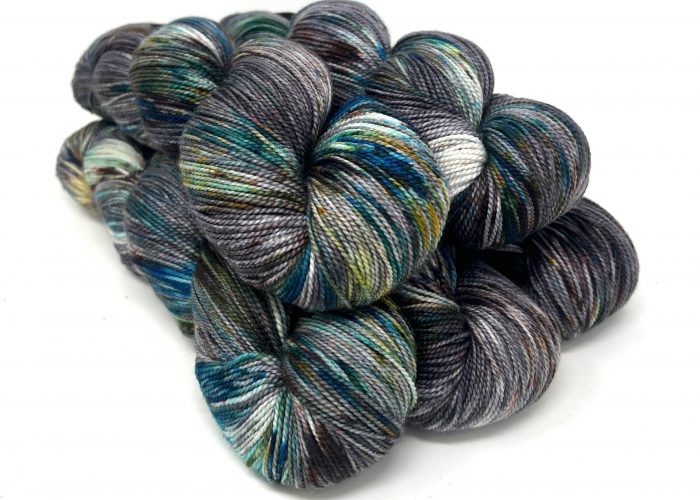 A pile of hand-dyed hanks of yarn with variegated colors of charcoal gray, navy, teal, olive green, and white, arranged on a plain white background.