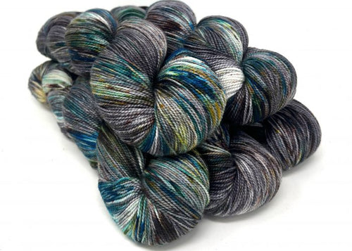 A pile of hand-dyed hanks of yarn with variegated colors of charcoal gray, navy, teal, olive green, and white, arranged on a plain white background.