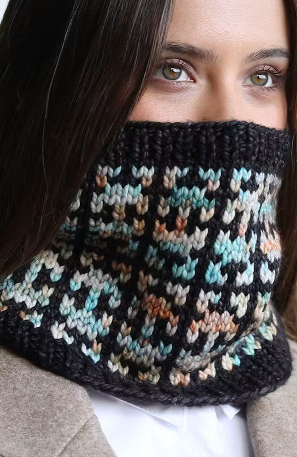 A close-up of a person wearing a chunky hand-knit cowl neck warmer. The neck warmer features a black base with a multicolor variegated pattern of light blue, beige, and orange.