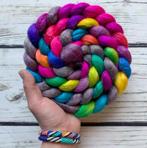 A hand holds a large, thick braided fiber roving made of many bright colors including pink, purple, blue, green, yellow, orange, and gray, against a whitewashed wood plank background.