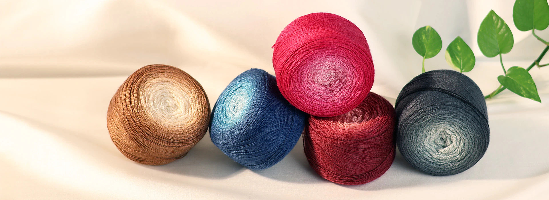 Four balls of variegated yarn in brown, blue, pink, and dark gray, arranged on a draped cream fabric background with a green leafy sprig in the corner.