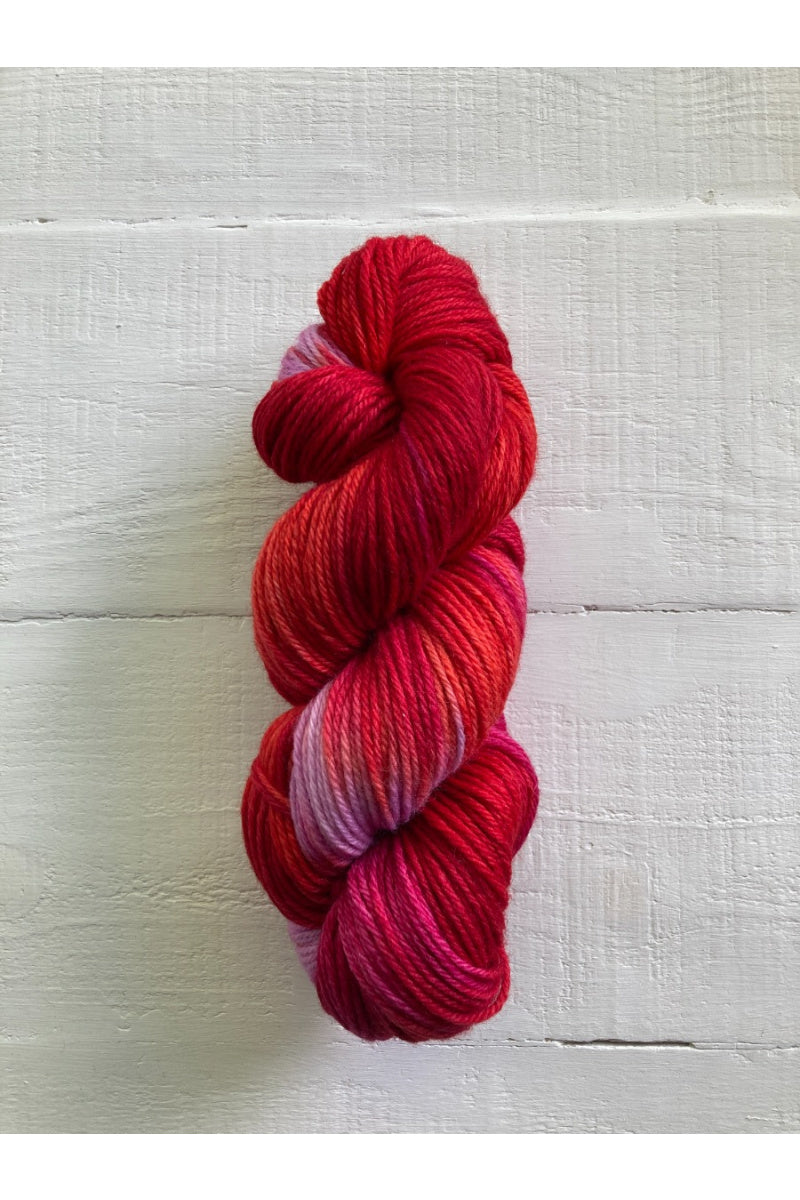 A single skein of variegated yarn in shades of red, pink, and purple, displayed vertically against a white painted wood plank background.