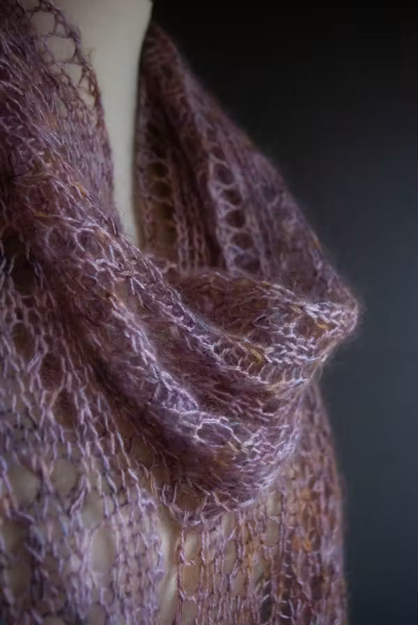 A close-up of a delicate, open-knit cowl scarf in a dusty mauve-purple color, draped on a mannequin against a dark background.