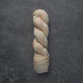 A single twisted skein of variegated yarn in a light oatmeal beige color with scattered orange, pink, and green flecks, displayed on a dark textured slate background.