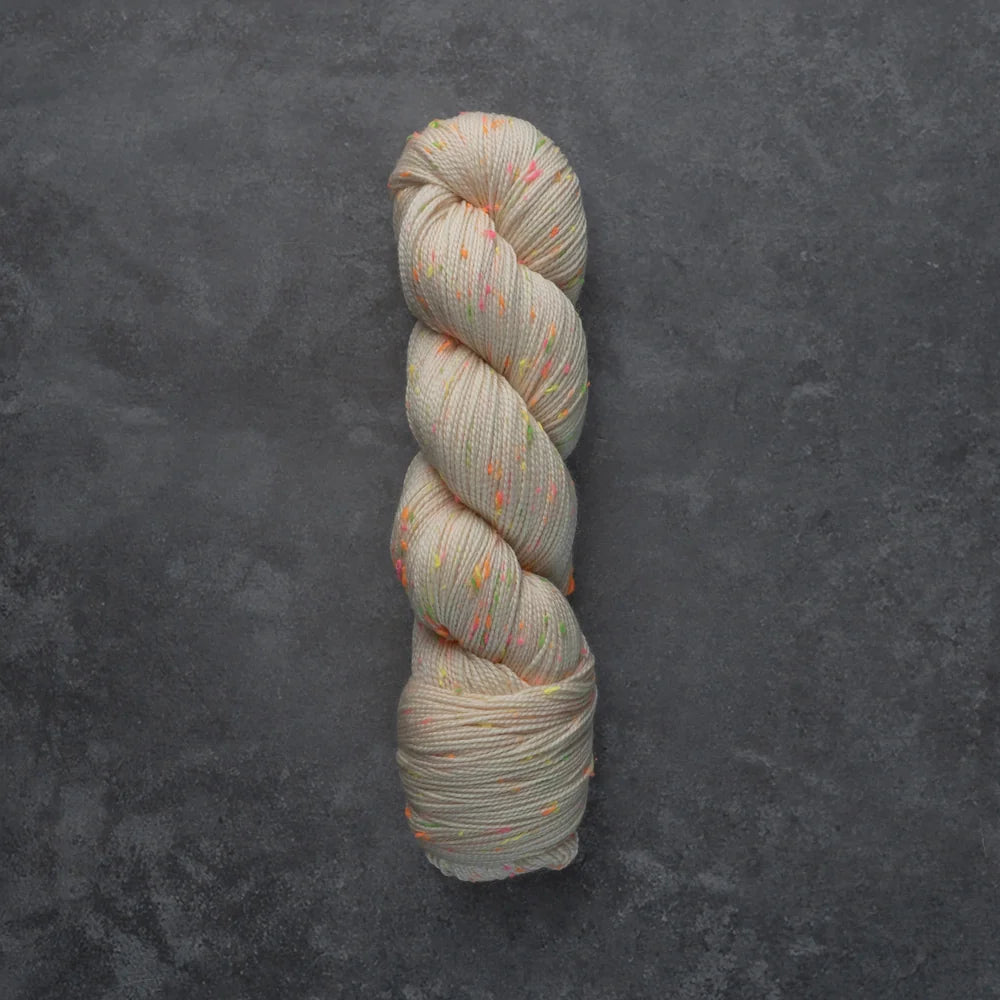 A single twisted skein of variegated yarn in a light oatmeal beige color with scattered orange, pink, and green flecks, displayed on a dark textured slate background.