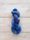 A twisted skein of variegated blue and gray yarn resting on a light-colored wooden plank surface.