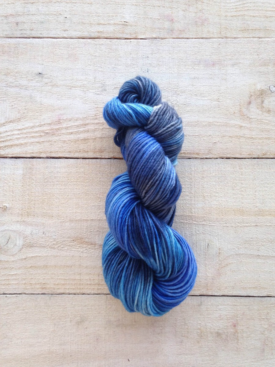 A twisted skein of variegated blue and gray yarn resting on a light-colored wooden plank surface.