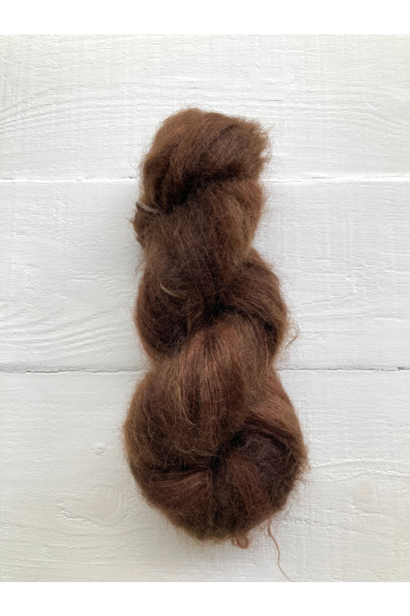 A bundle of medium brown curly hair extensions or a hair swatch, displayed vertically against a white painted wood plank background.