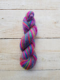 A twisted skein of vibrant, multicolored yarn with pink, blue, purple, green, and yellow strands, displayed vertically on a light-colored wooden plank background.