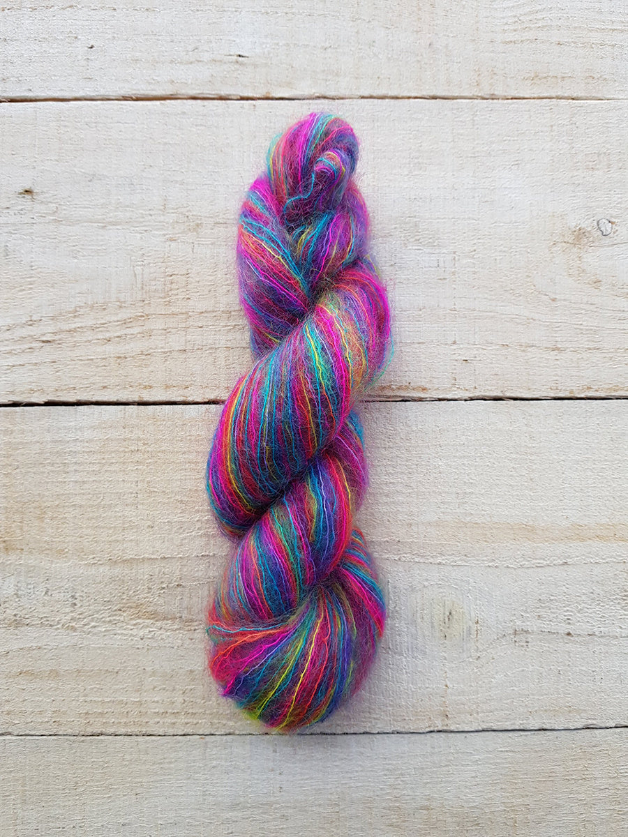 A twisted skein of vibrant, multicolored yarn with pink, blue, purple, green, and yellow strands, displayed vertically on a light-colored wooden plank background.