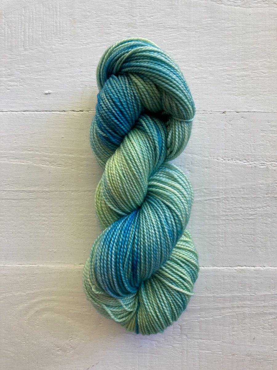 A single skein of variegated yarn in shades of teal, blue, and mint green, displayed on a white painted wood background.